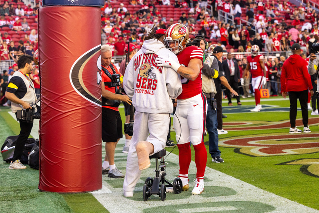 A football player in San Francisco 49ers gear hugs a man on a walker wearing a 49ers hoodie and a cast on his foot by the end zone.